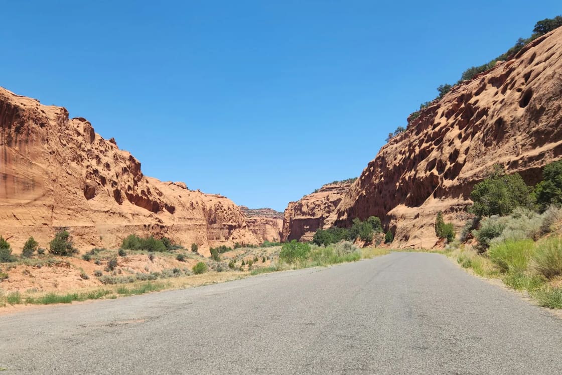 A paved two-lane road running between red sandstone canyon walls in Grand Staircase-Escalante country under a clear blue sky