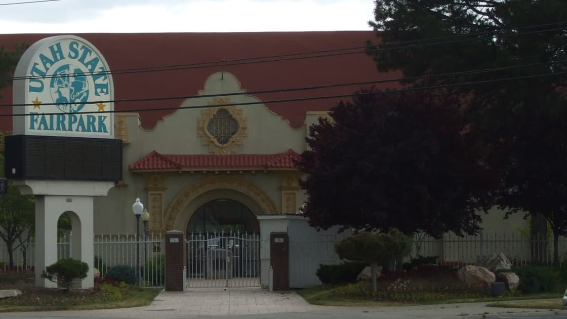 The Spanish-style entrance building of the Utah State Fairpark in Salt Lake City, with a red tile roof, an ornate gable, an arched gated entry and the Fairpark's round sign, framed by trees