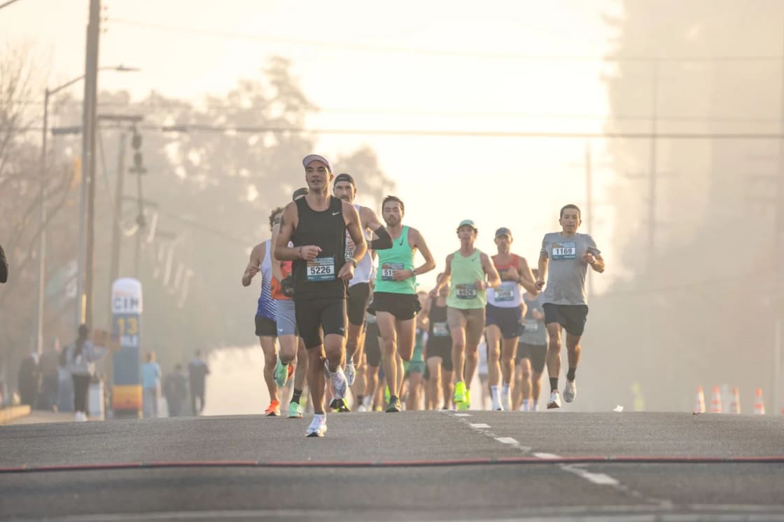 A pack of marathon runners wearing race bibs comes down a wide road at sunrise in hazy golden light, a mile marker and roadside cones behind them