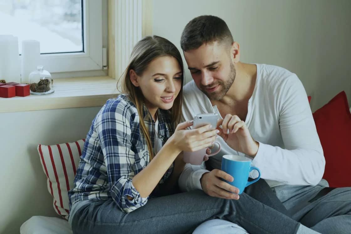 A young couple sits close together on a window seat, coffee mug in hand, looking over something on her phone.