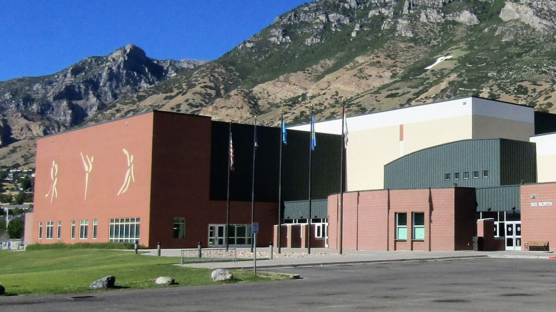 Peaks Ice Arena in Provo on a clear summer day, silhouettes of skaters on its brick end wall and flags out front, beneath the steep face of the Wasatch Range