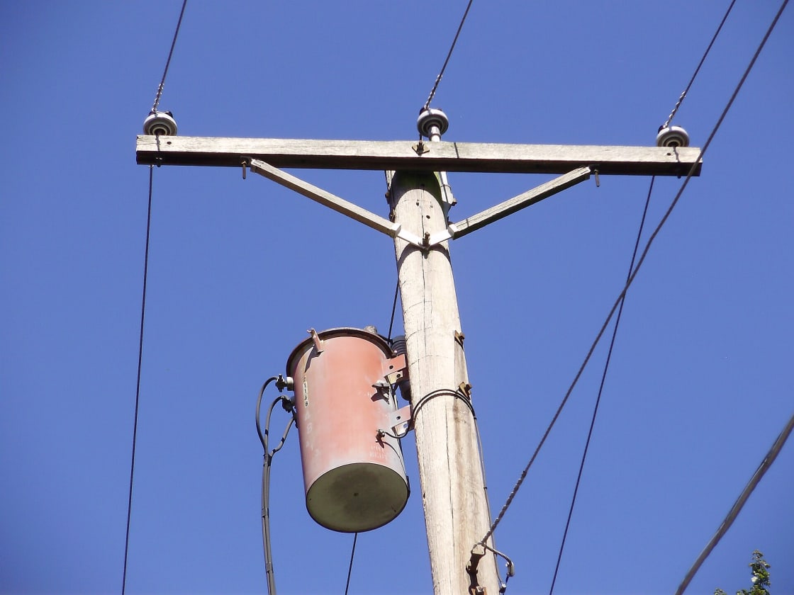 A wooden utility pole carrying a cylindrical distribution transformer and crossarm insulators against a clear blue sky