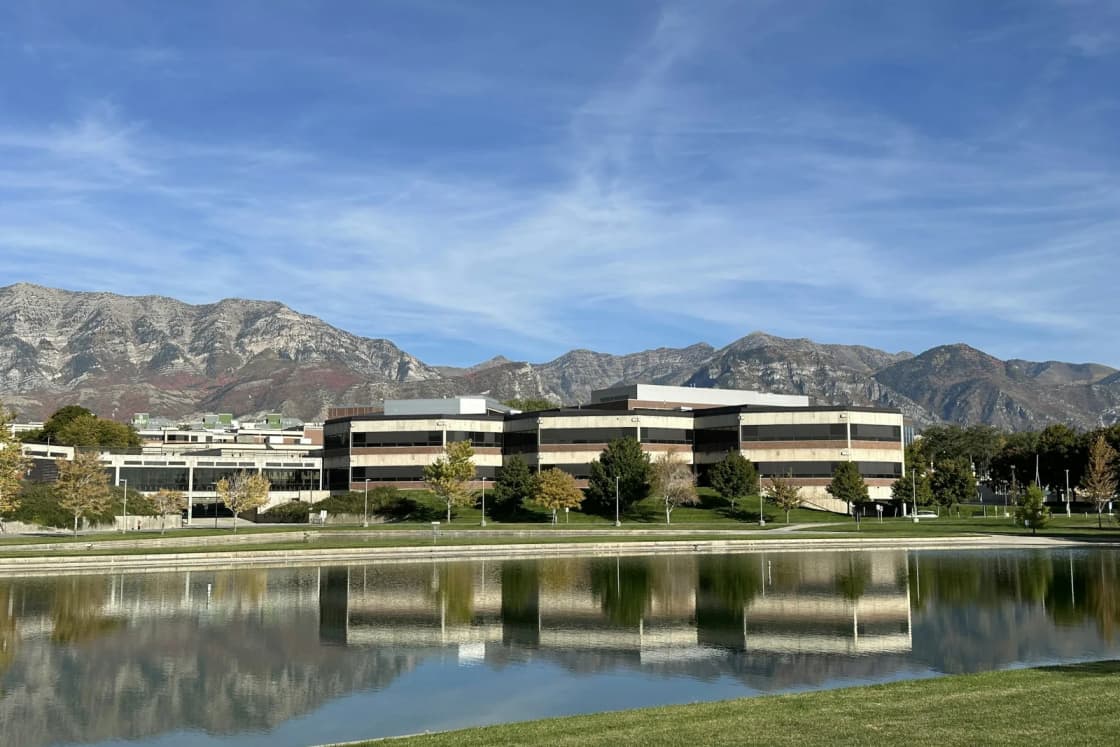 Low campus office buildings in Orem reflected in a still pond, the Wasatch Front's peaks rising behind them under a blue sky
