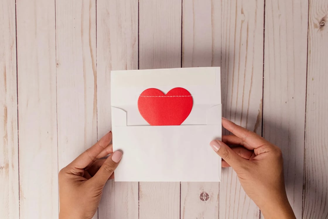Two hands hold a plain white envelope with a cut-paper red heart tucked inside, set against whitewashed wood boards.