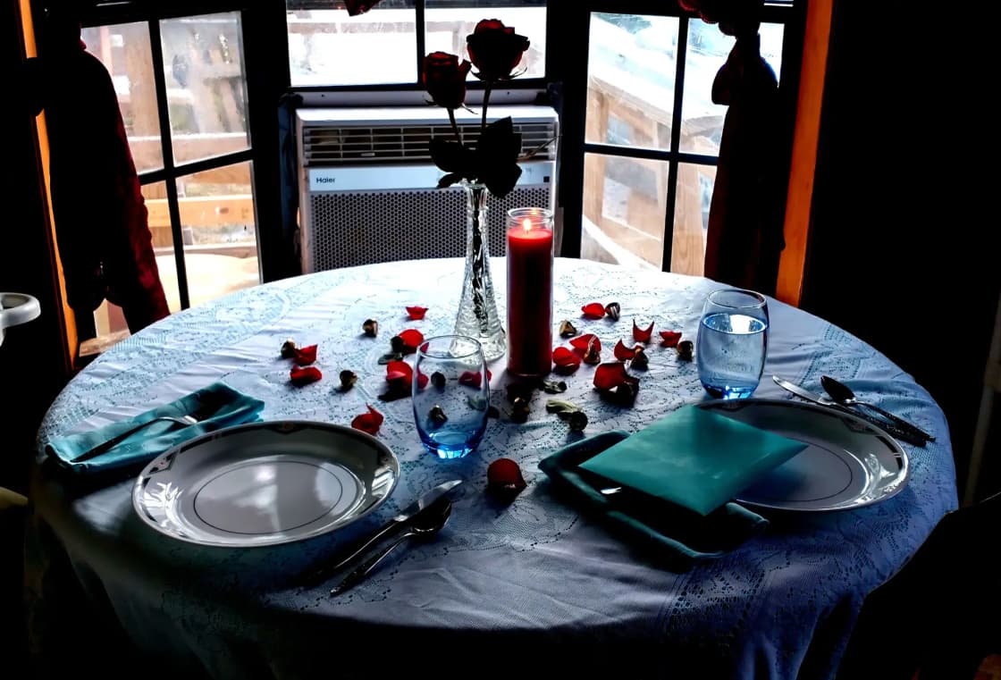 A restaurant table set for two beneath tall windows, a single red candle burning between the plates and rose petals scattered across the tablecloth