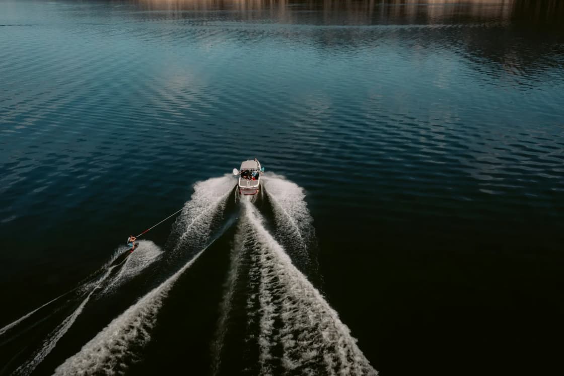 An overhead view of a motorboat towing a skier across dark open water, its white wake spreading in a long V behind