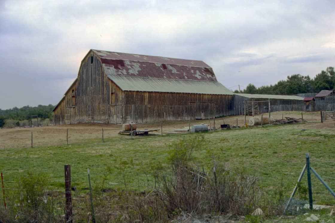 A large weathered wooden barn with a rusty gambrel roof stands beyond a green pasture and fence posts in Utah.