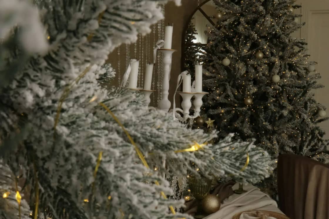 Frosted Christmas trees with warm string lights beside a long table set with plates and gold accents for a holiday dinner