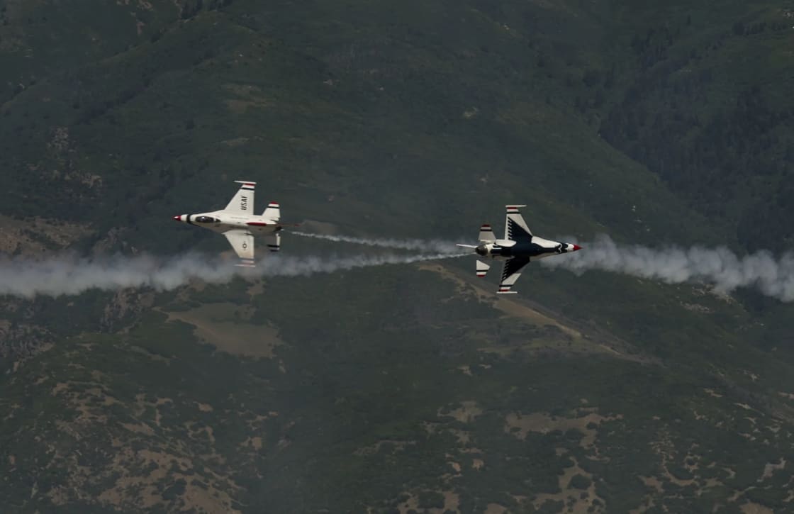 Two U.S. Air Force Thunderbirds F-16 jets in white, red and blue flying in formation with white smoke trailing behind them, low over dark, forested mountain slopes