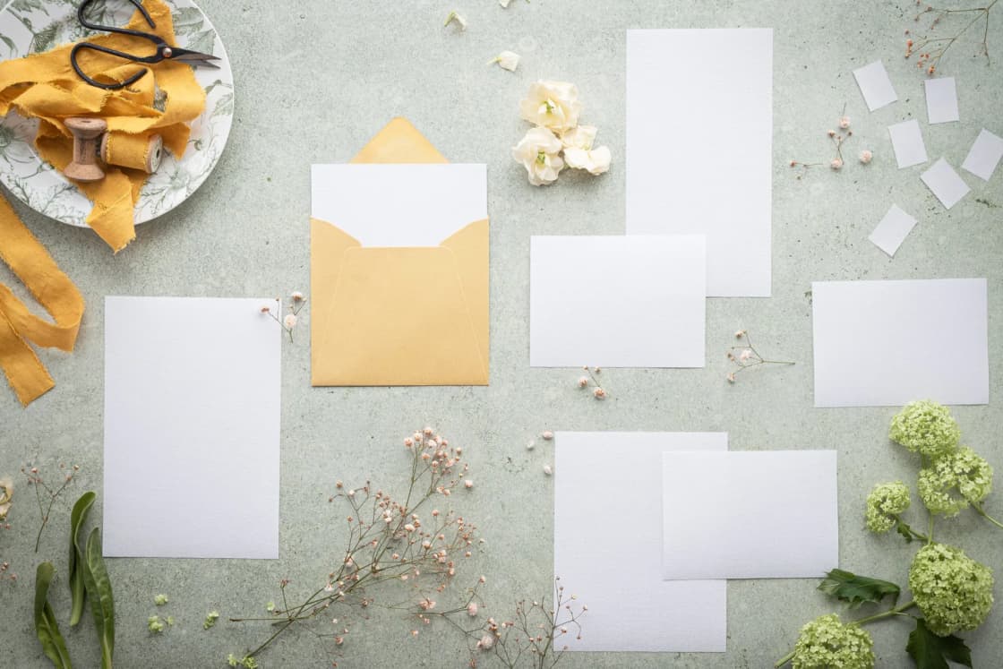 Blank invitation cards, envelopes and ribbon laid out with small flowers on a gray table.