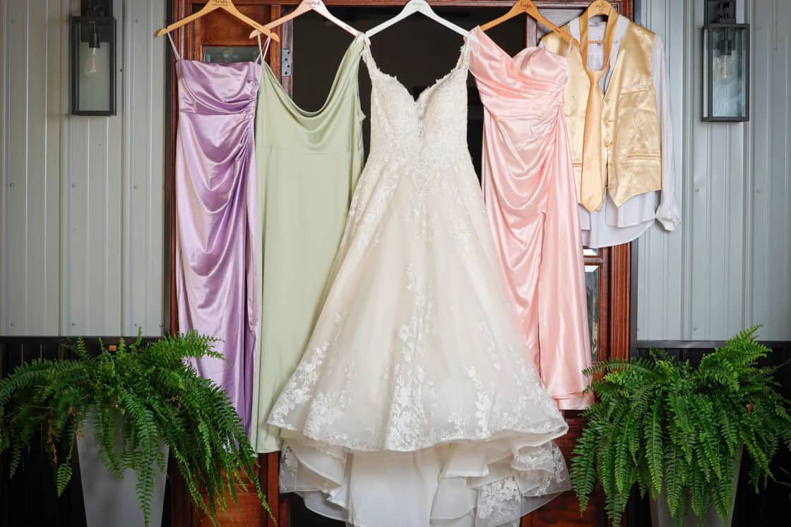 A white wedding gown and colored bridesmaid dresses hanging in a doorway between potted ferns.