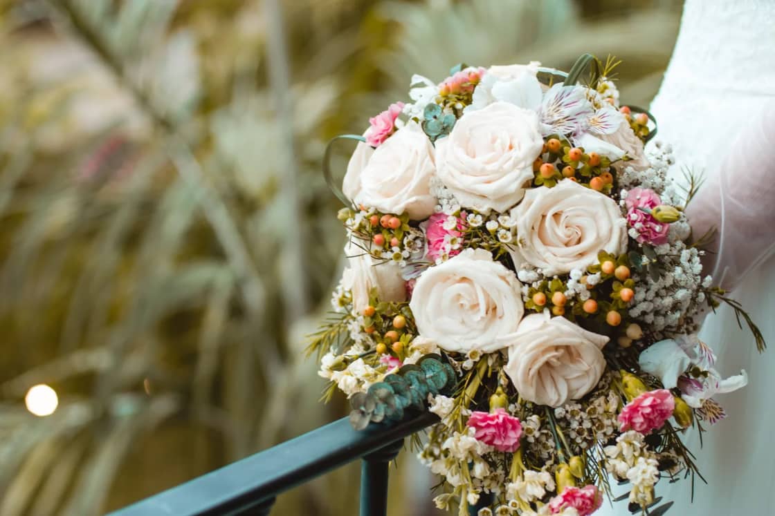 A bridal bouquet of cream roses and dried accents held against a soft, sunny background.