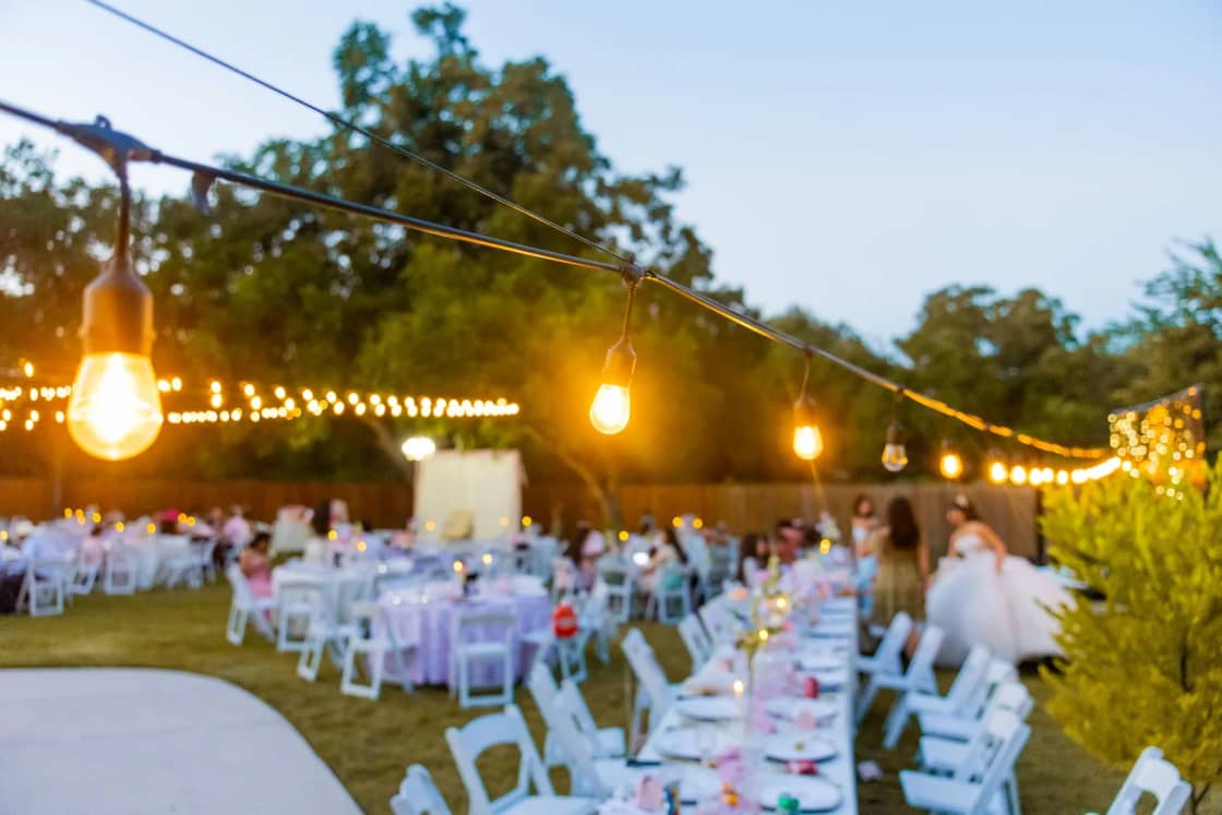 Guests at long outdoor reception tables under strings of glowing lights at dusk.