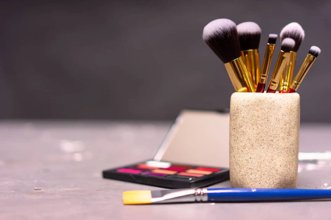 Makeup brushes standing in a cup beside an eyeshadow palette.