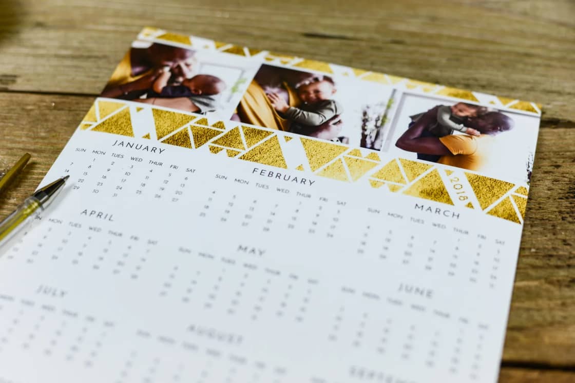 A printed year-at-a-glance calendar with a gold geometric border and family photos lies on a wooden table beside a gold pen.