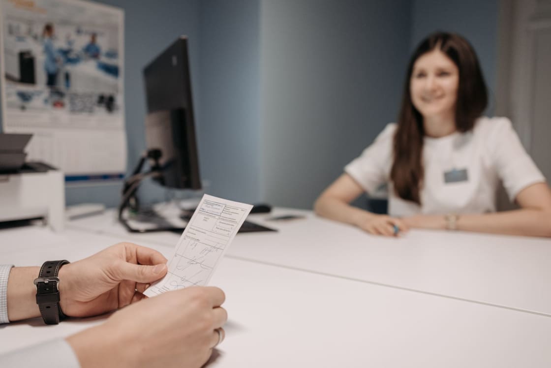 A clinician in white sitting behind a desk in a bright consultation room, smiling toward a patient in the foreground who is holding a small printed medical form, a monitor and printer on the desk behind