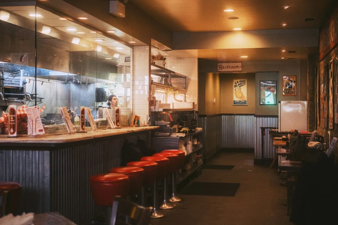 An empty diner interior with a row of red-topped chrome stools at a wood-paneled counter, warm pendant lights, and a window into the kitchen