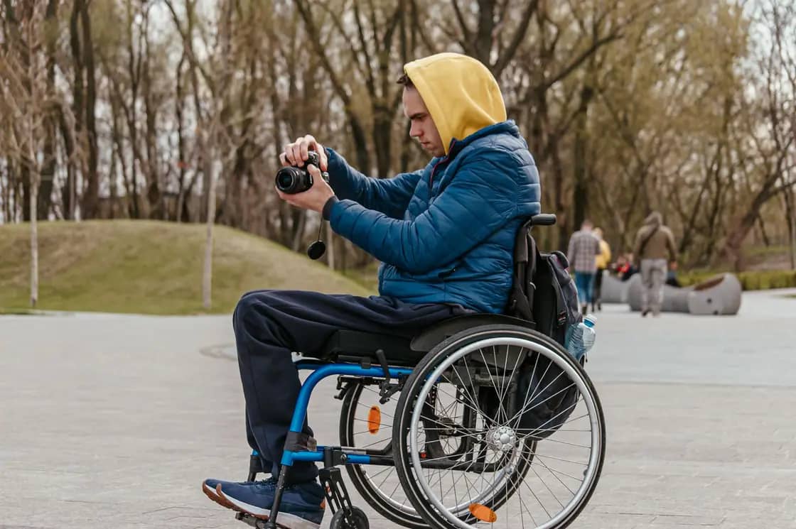 A man in a manual wheelchair taking a photograph on a wide paved path in a park
