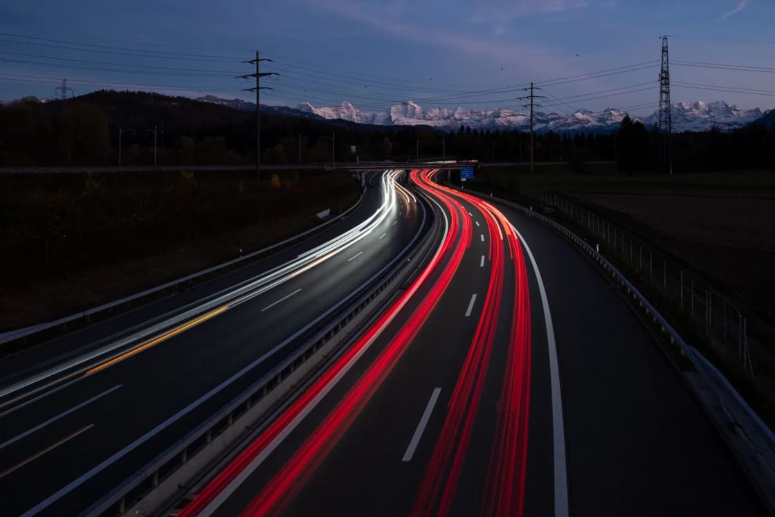 Red and white light trails streak along a curving freeway at dusk with snow-capped mountains on the horizon