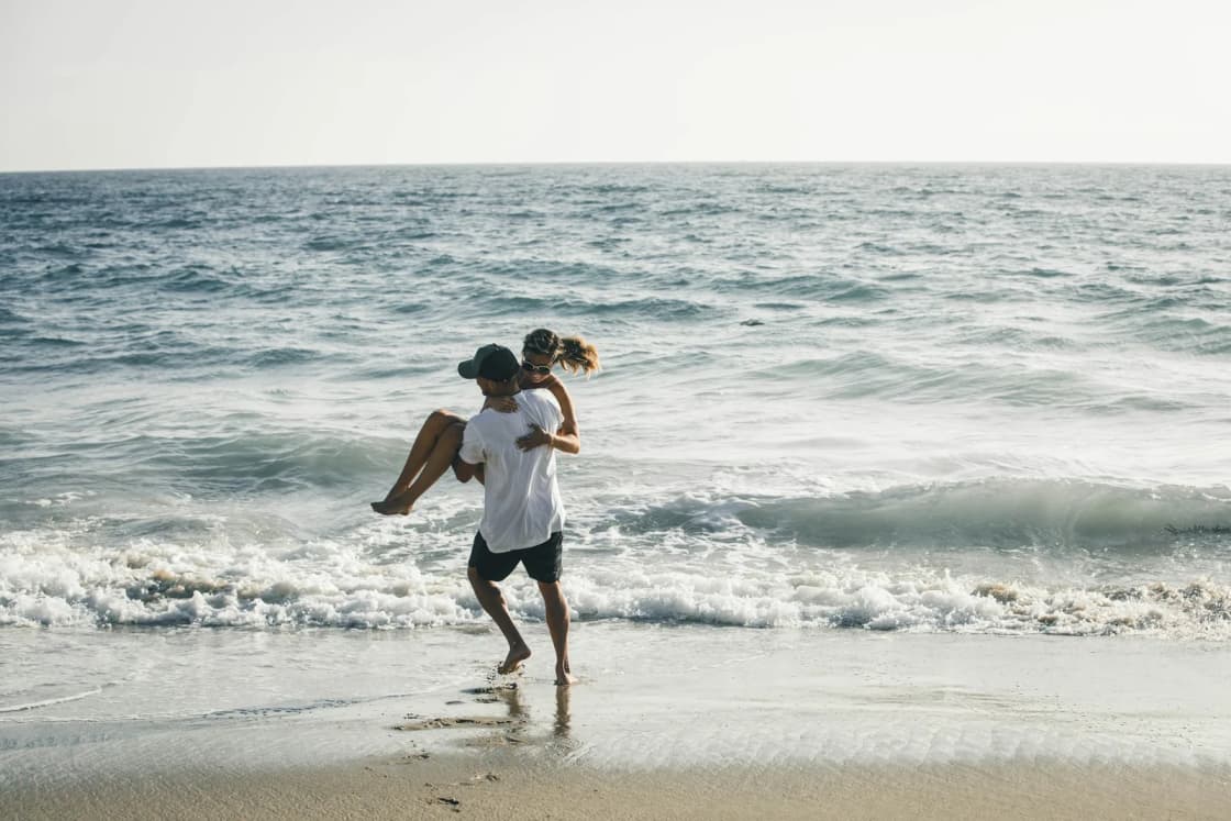 A man carries his laughing partner through the surf on a wide sandy beach as waves roll in under a hazy sky.