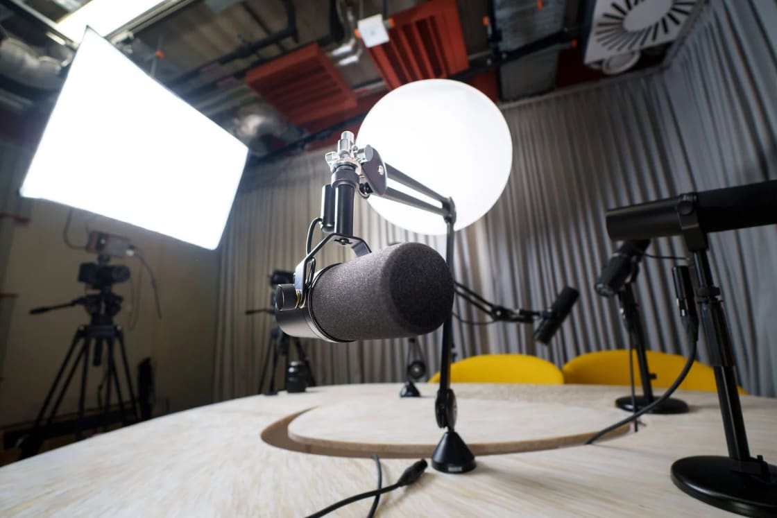 Microphones on boom arms around a round table in a studio lit by softbox lights, with a video camera on a tripod behind