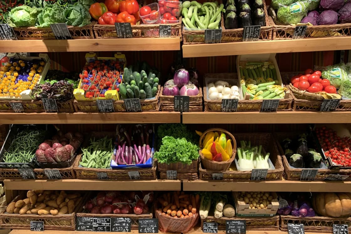 Rows of fresh vegetables fill wicker baskets on wooden market shelves, from red tomatoes on top to potatoes and carrots at the bottom.