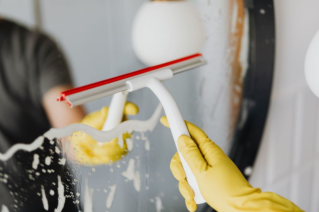 A yellow-gloved hand drawing a squeegee down soapy glass, leaving a clear stripe behind the blade