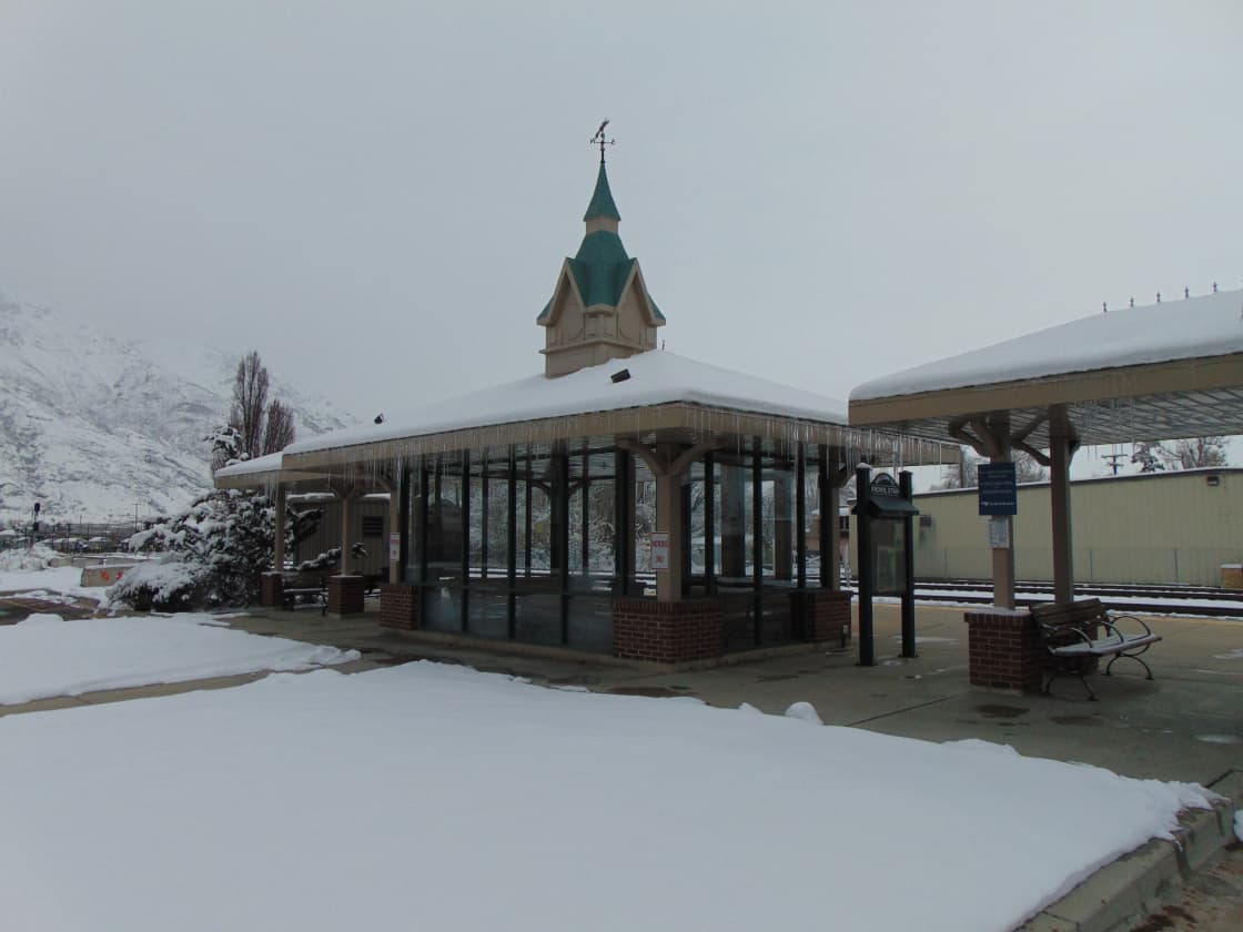 The snow-covered Provo train station under a flat gray sky, icicles hanging from its glass shelter and the platform and benches completely empty