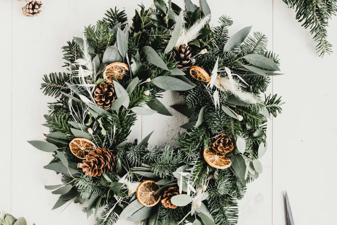 A homemade evergreen wreath studded with pine cones and dried orange slices lies on a white table beside cut fir boughs and scissors.