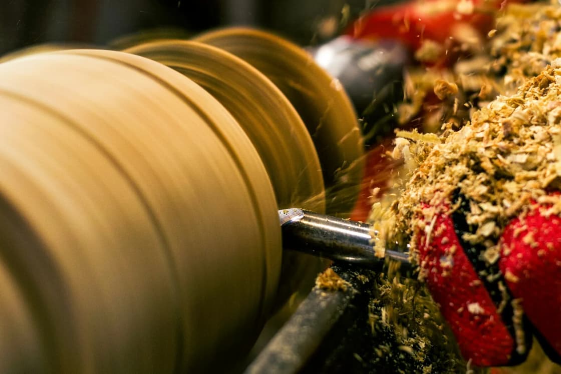Wood shavings fly from a spinning blank on a lathe as a turning tool cuts into it, shown close up in warm shop light.