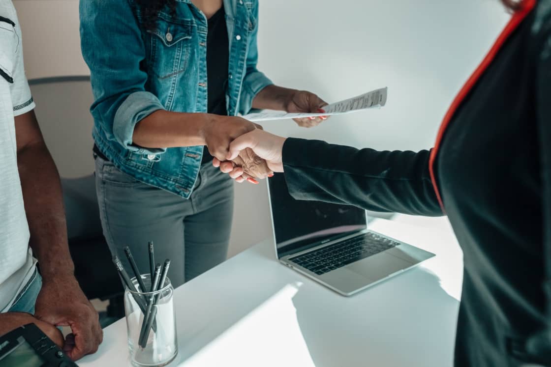 Two people shaking hands across a white desk, one holding a printed document, with a laptop and a jar of pencils beside them