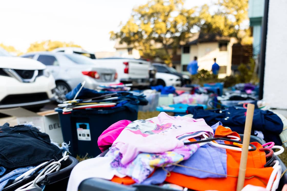 Clothing and goods piled on tables at an outdoor sale with parked cars behind