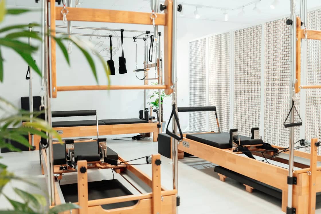A bright Pilates studio with wooden reformer and Cadillac tower machines lined up on a white floor beside a potted palm