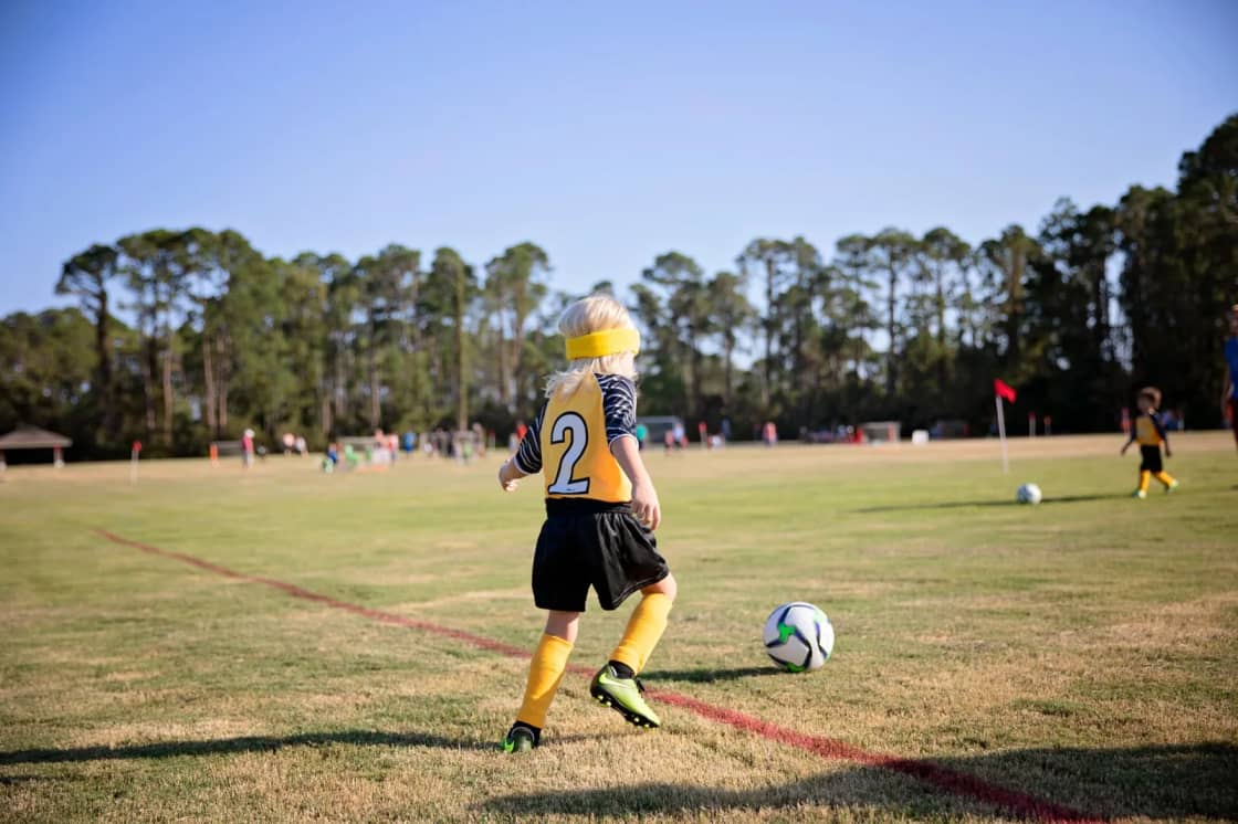 A young player in a yellow number-two jersey and black shorts runs with a soccer ball along a white sideline on a grass field, with tall trees and a few spectators in the distance