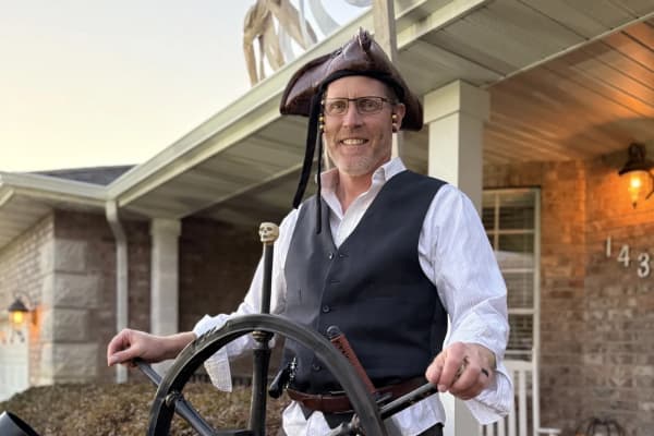 Gary Gibb, smiling, in a leather tricorn hat, white shirt and dark vest, holding the wheel of his pirate ship on his front porch