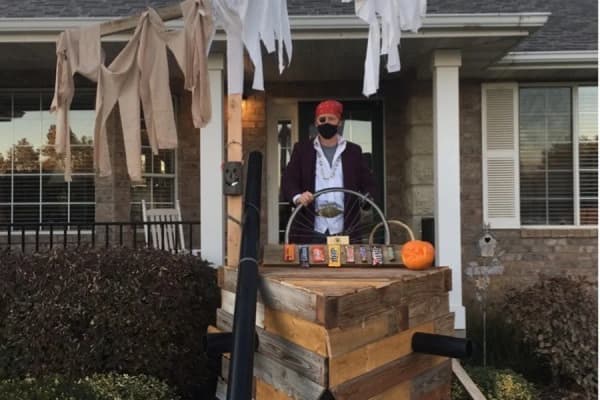Gary Gibb in a mask and red bandana at the wheel of a small wooden pirate ship built on his front porch in 2020, with a black pipe cannon on its side