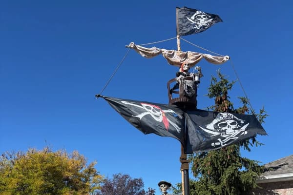 A tall pirate mast in daylight with a crow's nest, a tattered sail and two Jolly Roger flags against a bright blue sky