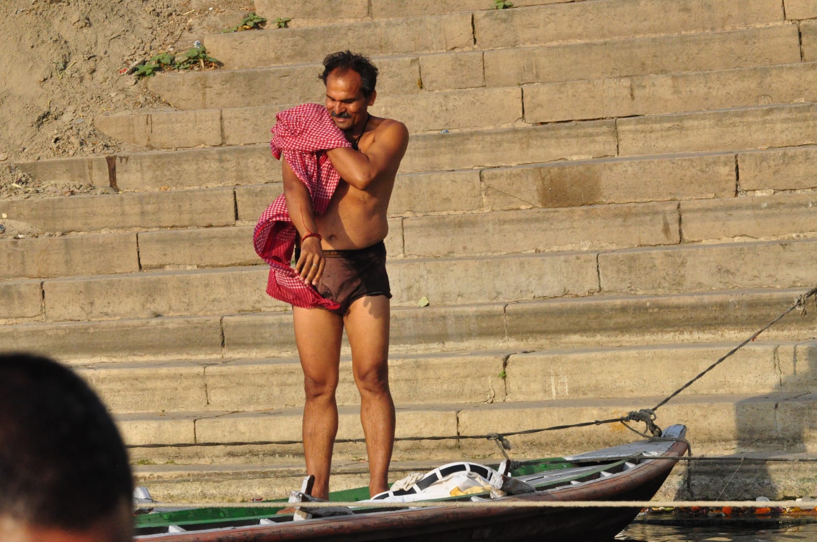 Pandu drying his shoulder with a red checked cloth beside a boat and stone steps.