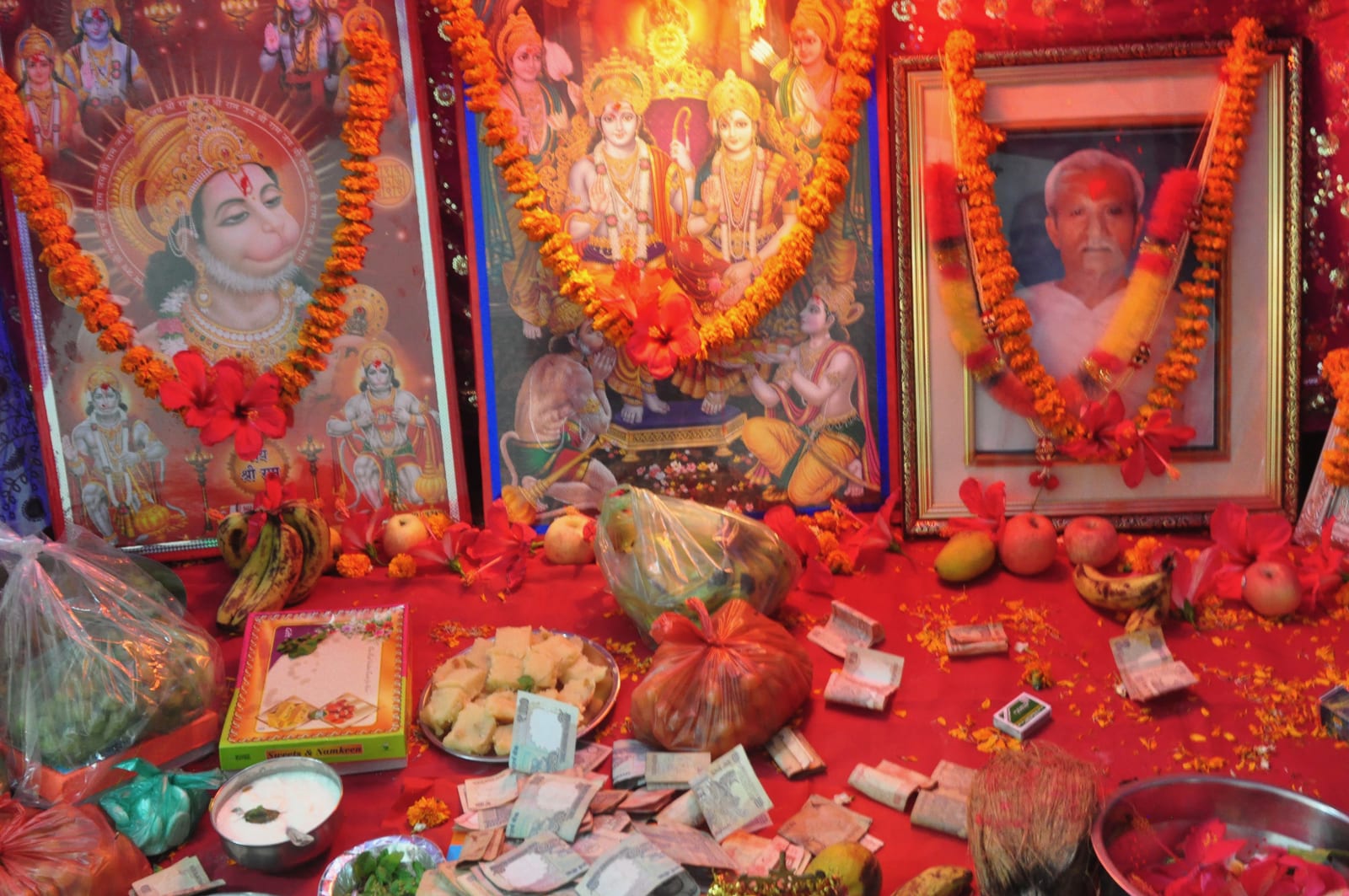 Garlanded religious pictures and a framed portrait above offerings, fruit and currency notes.