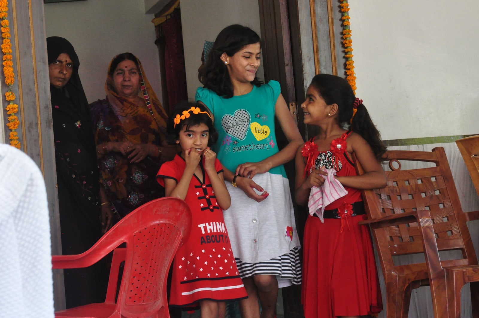 Three girls standing beside chairs in the doorway, with women seated behind them.