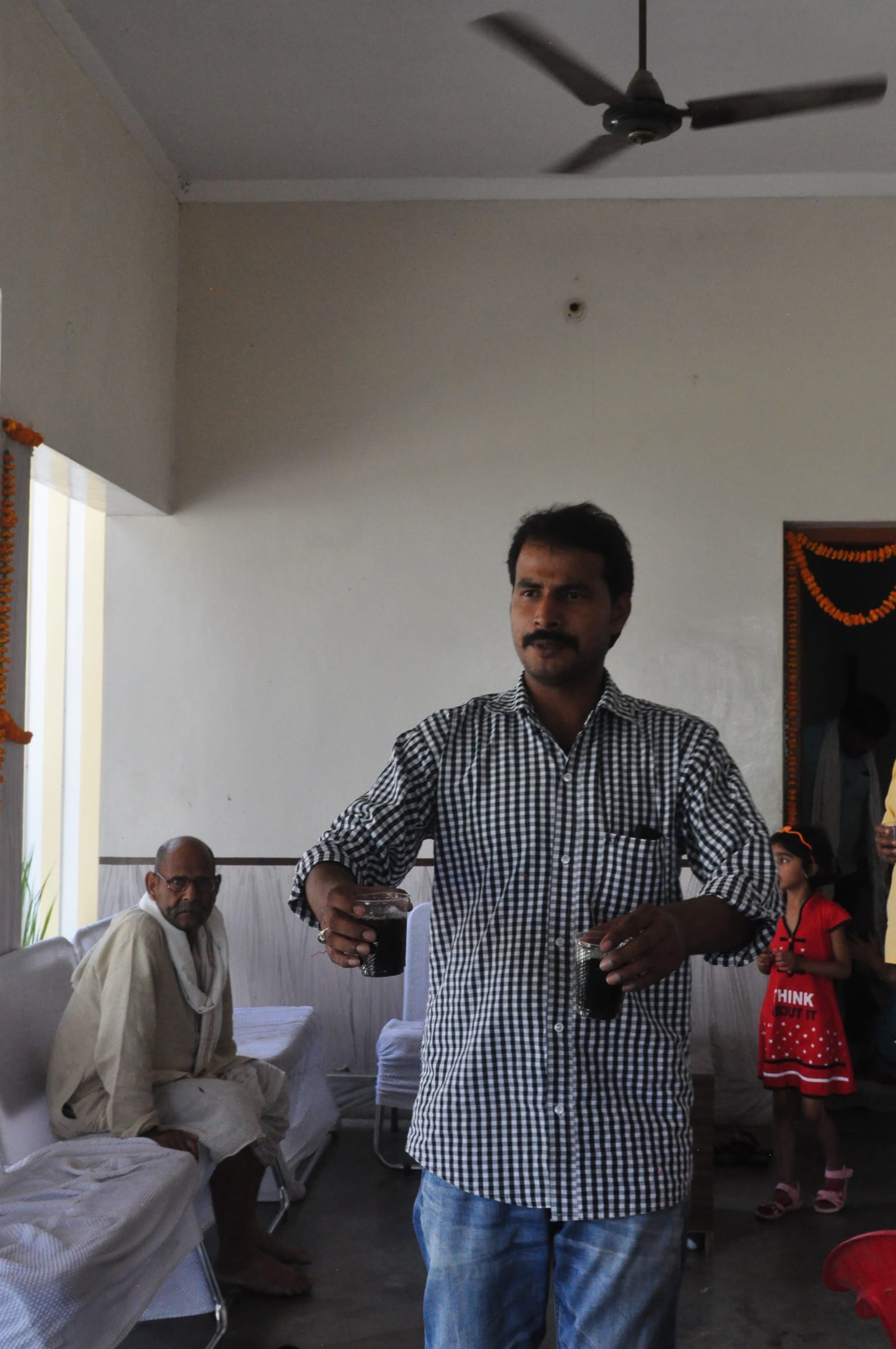 A man in a checked shirt standing and gesturing inside the room, with seated participants behind him.