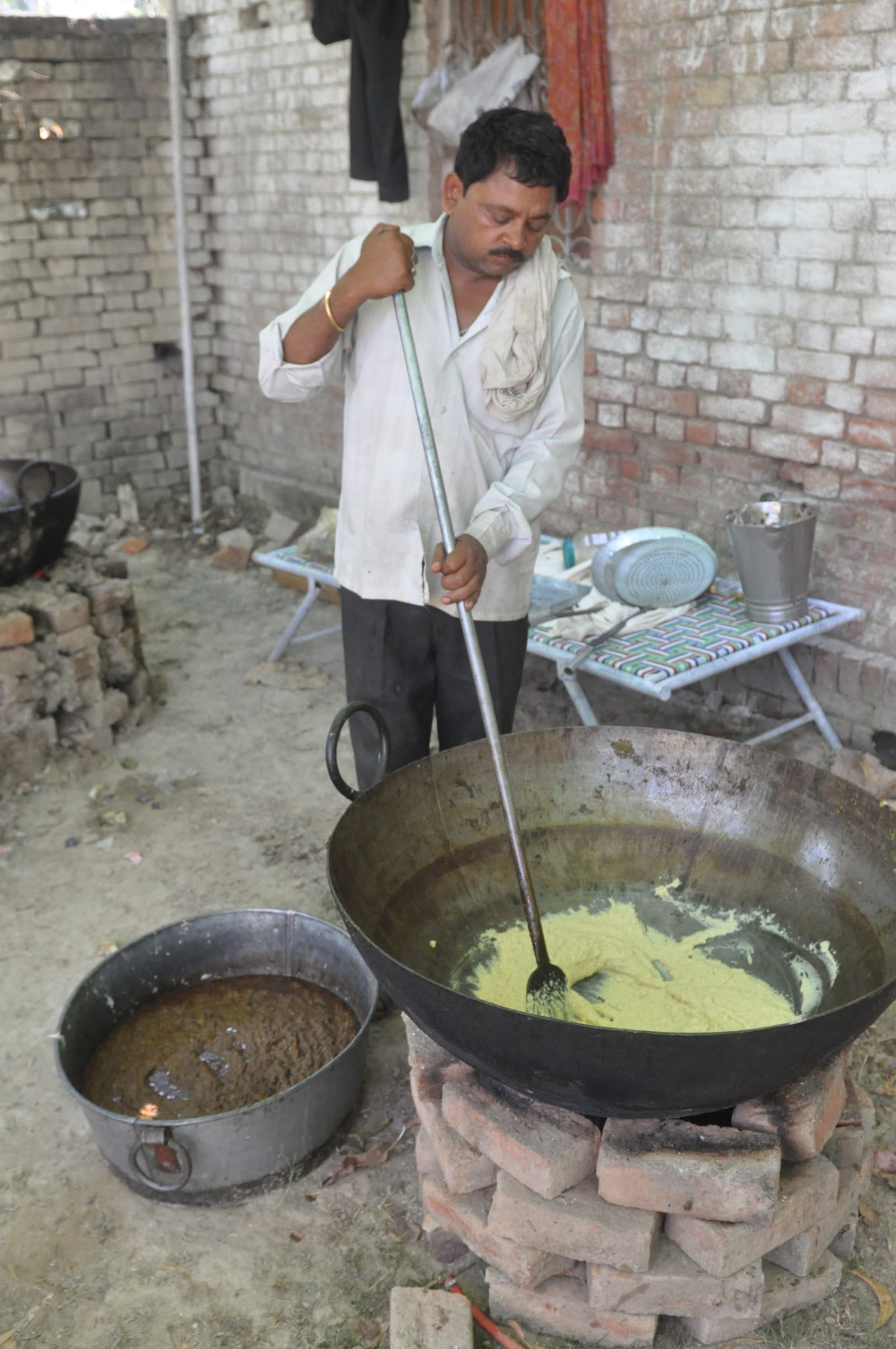A man using a long utensil to stir a large cooking vessel set on a brick stove.