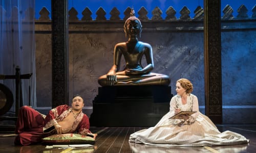 Golygfa o King and I, yn cynnwys dyn mewn gwisg dwyreinol a menyw yn eistedd ar y llawr yn gwisgo ffrog, ger cerflun mawr.