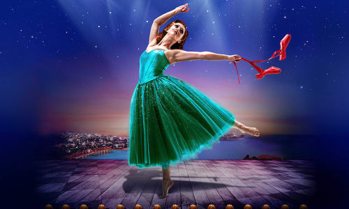 Ballerina in green dress dancing under the spotlight with a pair of red shoes in her hand.
