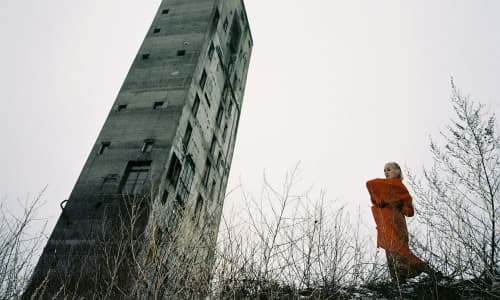 Cate Le Bon stood outside in a red coat next next to a grey, derelict tower.