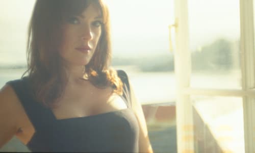 A woman with brown hair wearing a strappy black dress stares at the camera