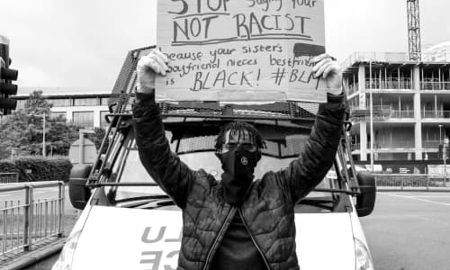 A young black man wearing a mask sits on a police van's bonnet holding up a protest banner