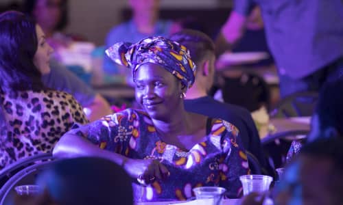 A smiling black woman sat down, wearing a colourful dress 