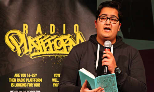 Awais from Radio Plattform holding a mic and poetry book at a spoken word performance at Wales Millennium Centre