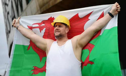 Yr actor Kai Owen yn gwisgo fest gwyn, yn dal fflag Cymru tu allan i Ganolfan Mileniwm Cymru